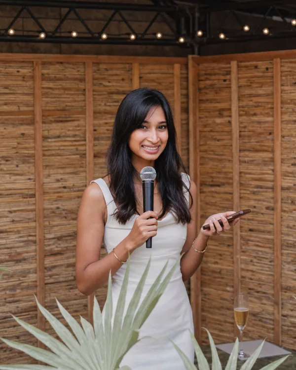 Rushmi holding a microphone, smiling to camera in front of a warm timber wall, string lights above.
